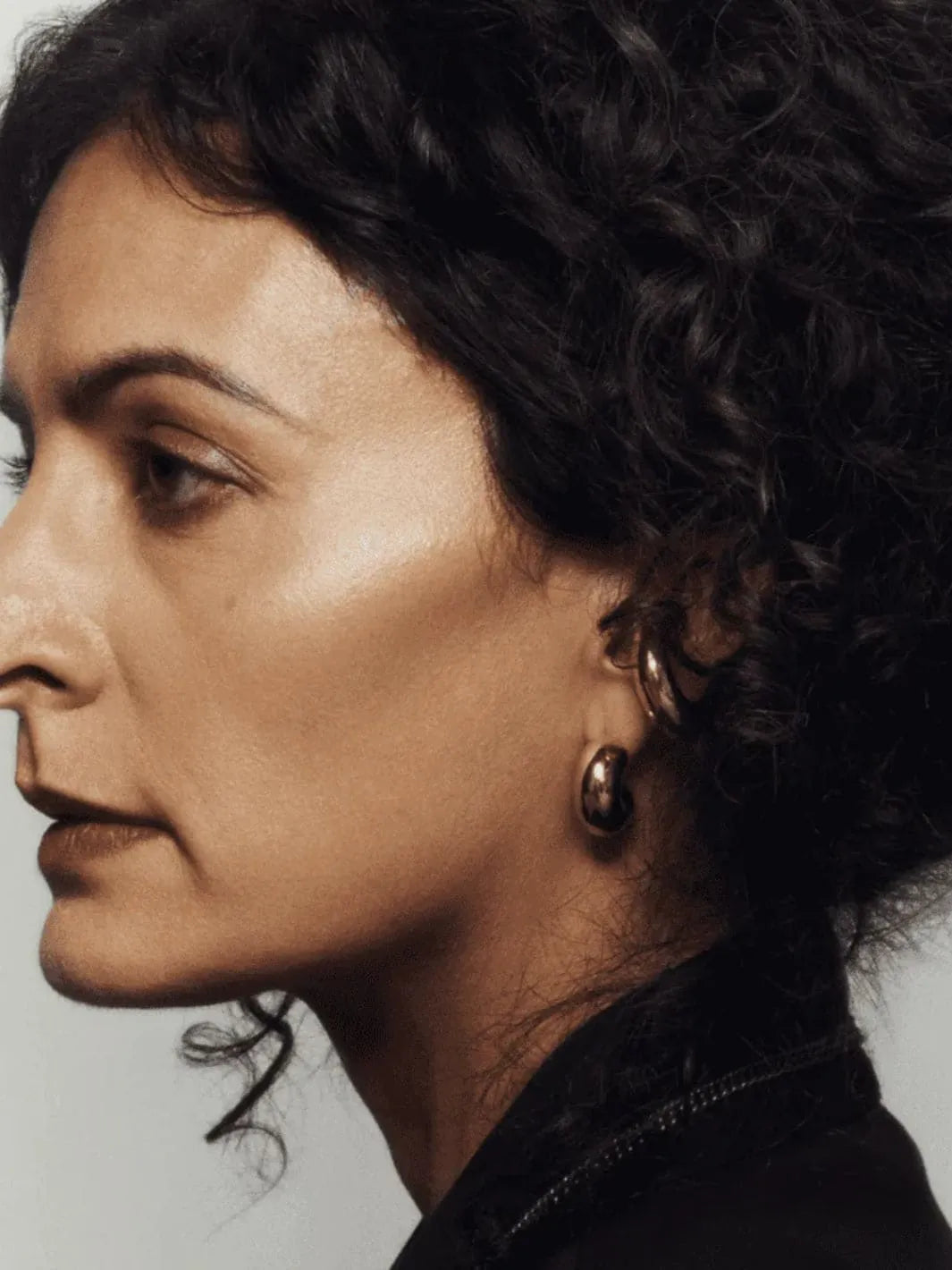 Close-up of a person with dark hair and earrings using 19/99 Beauty Cream Contour Stick in 'Tea' against a neutral background | Twentyseven Toronto