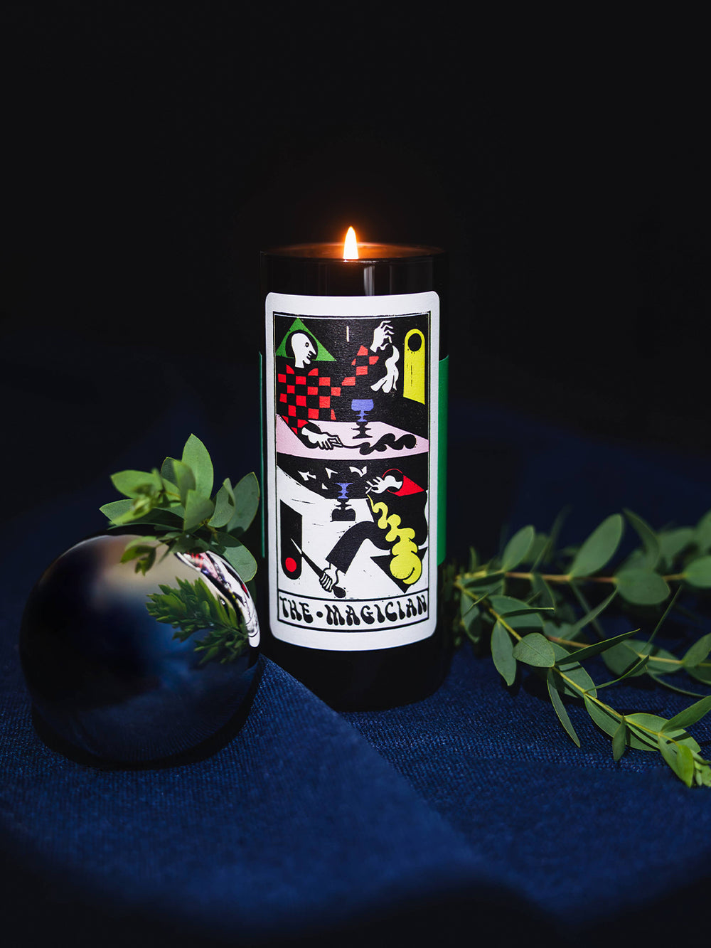 Lit 54 Celsius The Magician Tarot Candle styled with eucalyptus leaves against a dark blue background | Twentyseven Toronto