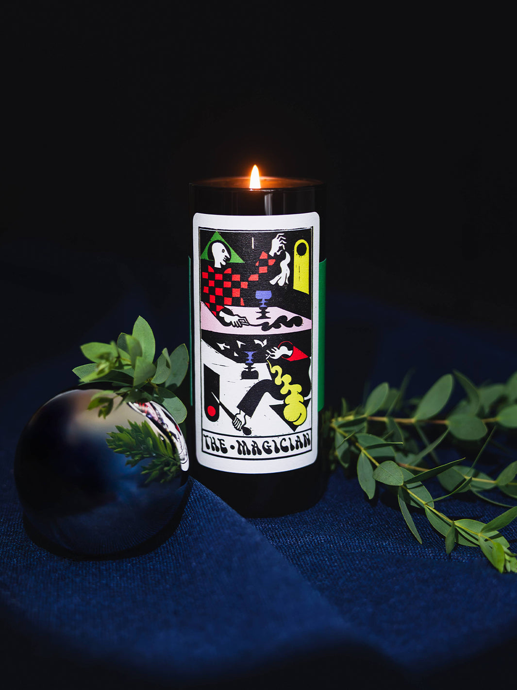 Lit 54 Celsius The Magician Tarot Candle styled with eucalyptus leaves against a dark blue background | Twentyseven Toronto