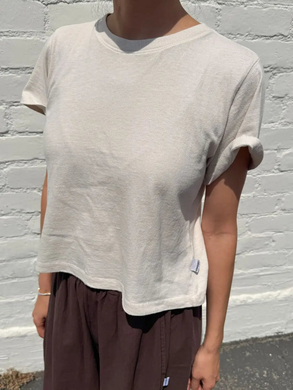 A close-up side view of a model wearing the Le Bon Shoppe Airy Darling Tee in Rice paired with dark brown trousers against a painted white brick wall | Twentyseven
