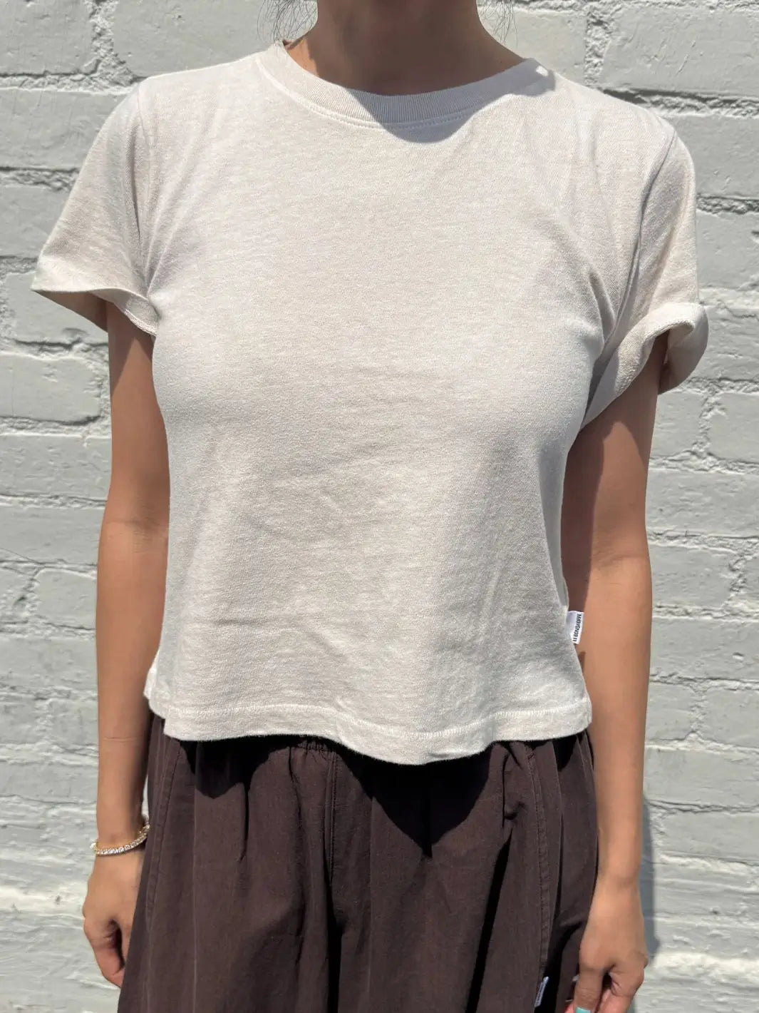 A front view shot of a model wearing the Le Bon Shoppe Airy Darling Tee in Rice standing against a white brick wall | Twentyseven