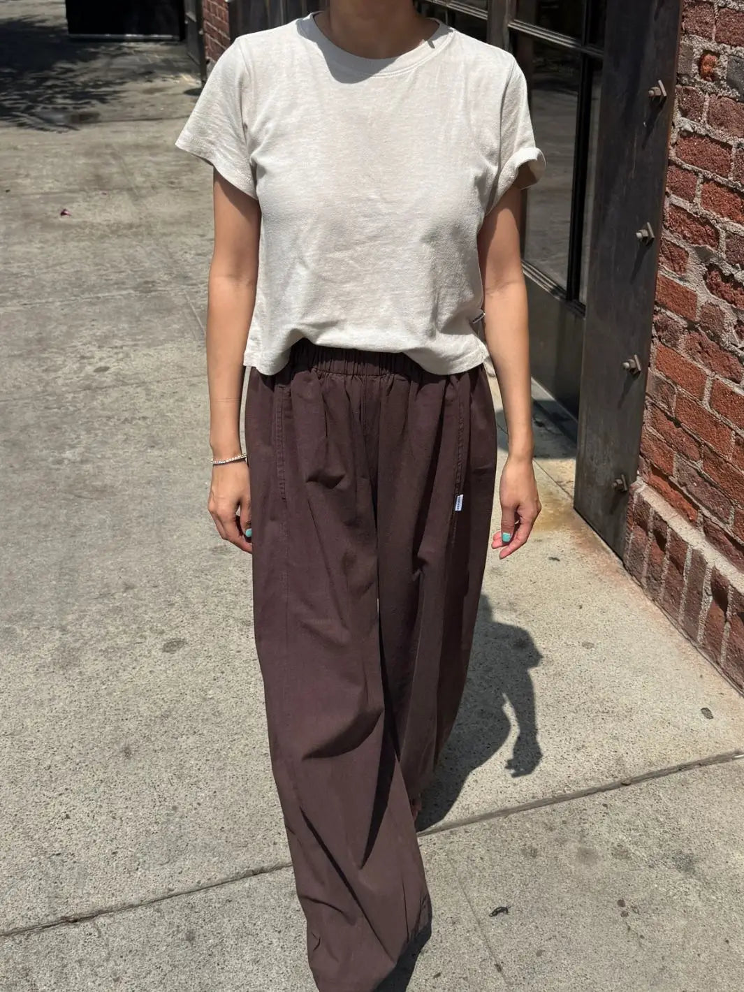 A full-length outdoor shot of a model walking down a sidewalk wearing the Le Bon Shoppe Airy Darling Tee in Rice with loose brown pants | Twentyseven