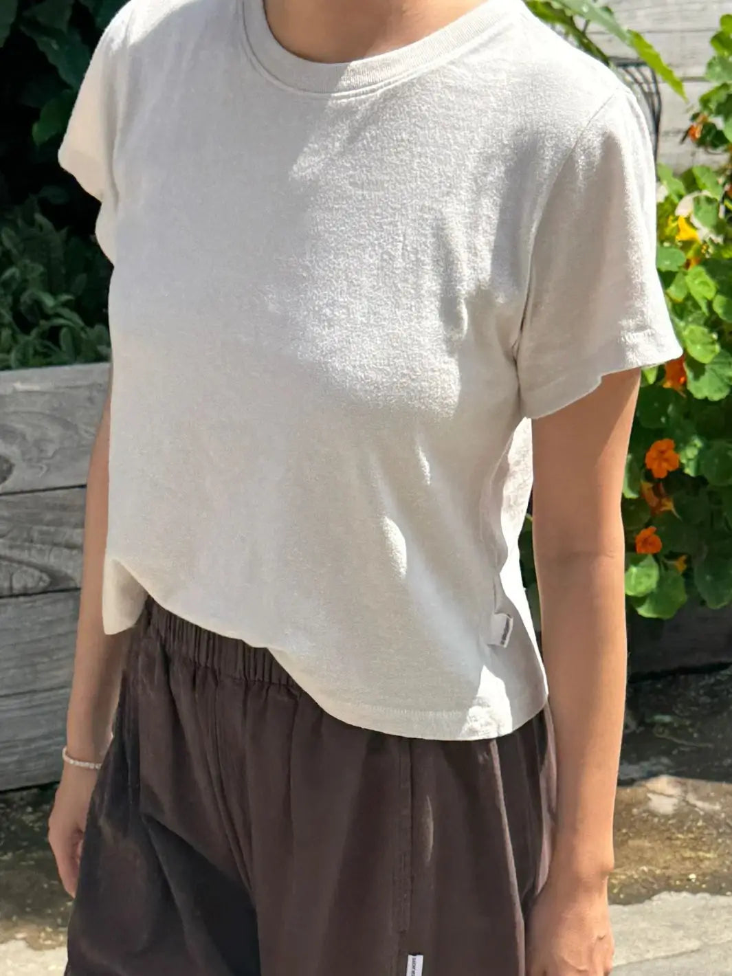 An outdoor close-up shot of a model wearing the Le Bon Shoppe Airy Darling Tee in Rice standing in natural sunlight near garden plants | Twentyseven
