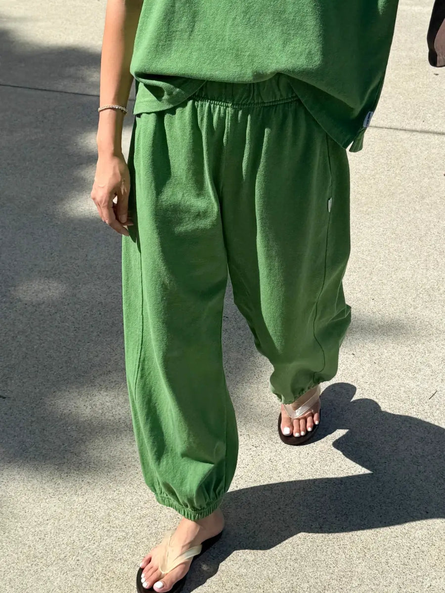 A walking shot of a model wearing the Le Bon Shoppe Balloon Pants in Asparagus styled with matching green top and sandals on a sunny pavement | Twentyseven