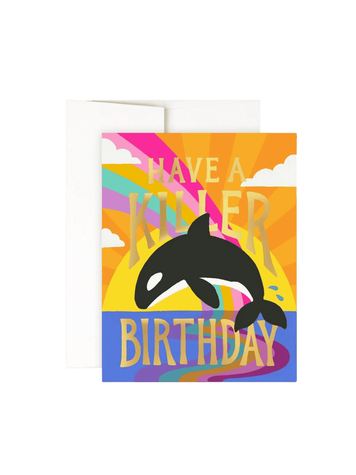 A studio product shot of the Idlewild Co. Birthday Greeting Card in Killer Birthday featuring an orca whale leaping in front of a colorful sunset with gold foil lettering, set against a plain white envelope | Twentyseven