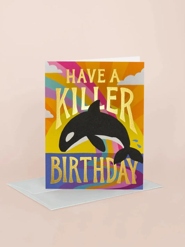 An angled lifestyle shot of the Idlewild Co. Birthday Greeting Card - Killer Birthday standing upright next to its white envelope against a soft beige background, highlighting the shiny gold foil text "Have a Killer Birthday" | Twentyseven