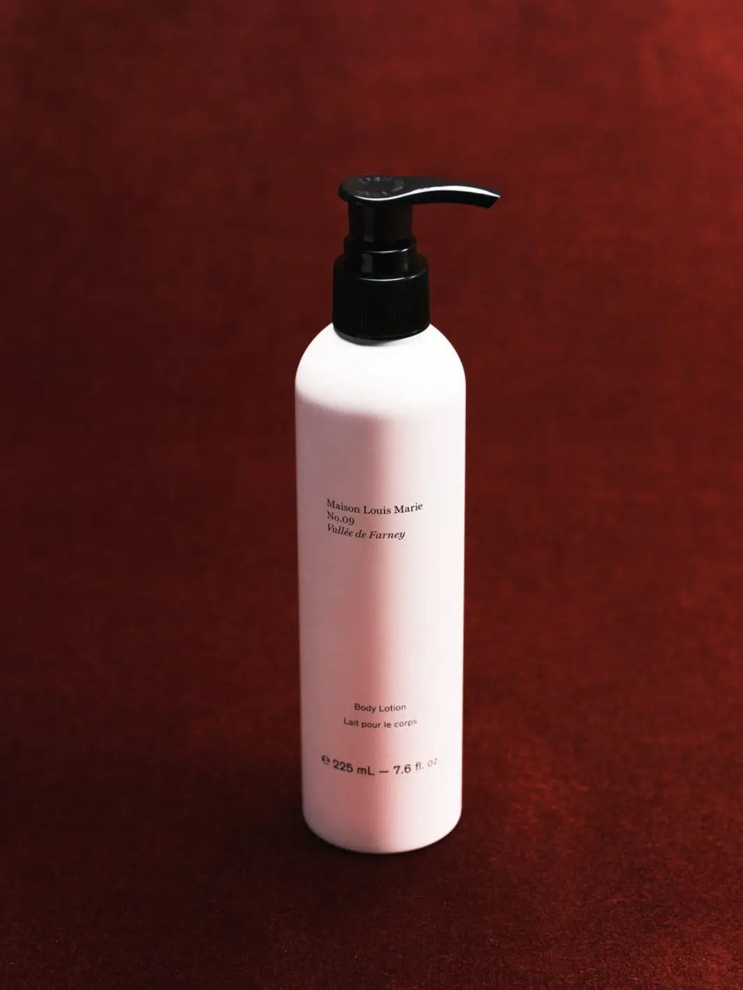 A 325 mL bottle of body lotion by Maison Louis Marie in Vallée de Farney, featuring a matte white bottle and a black pump dispenser, set against a dark textured red background | Twentyseven