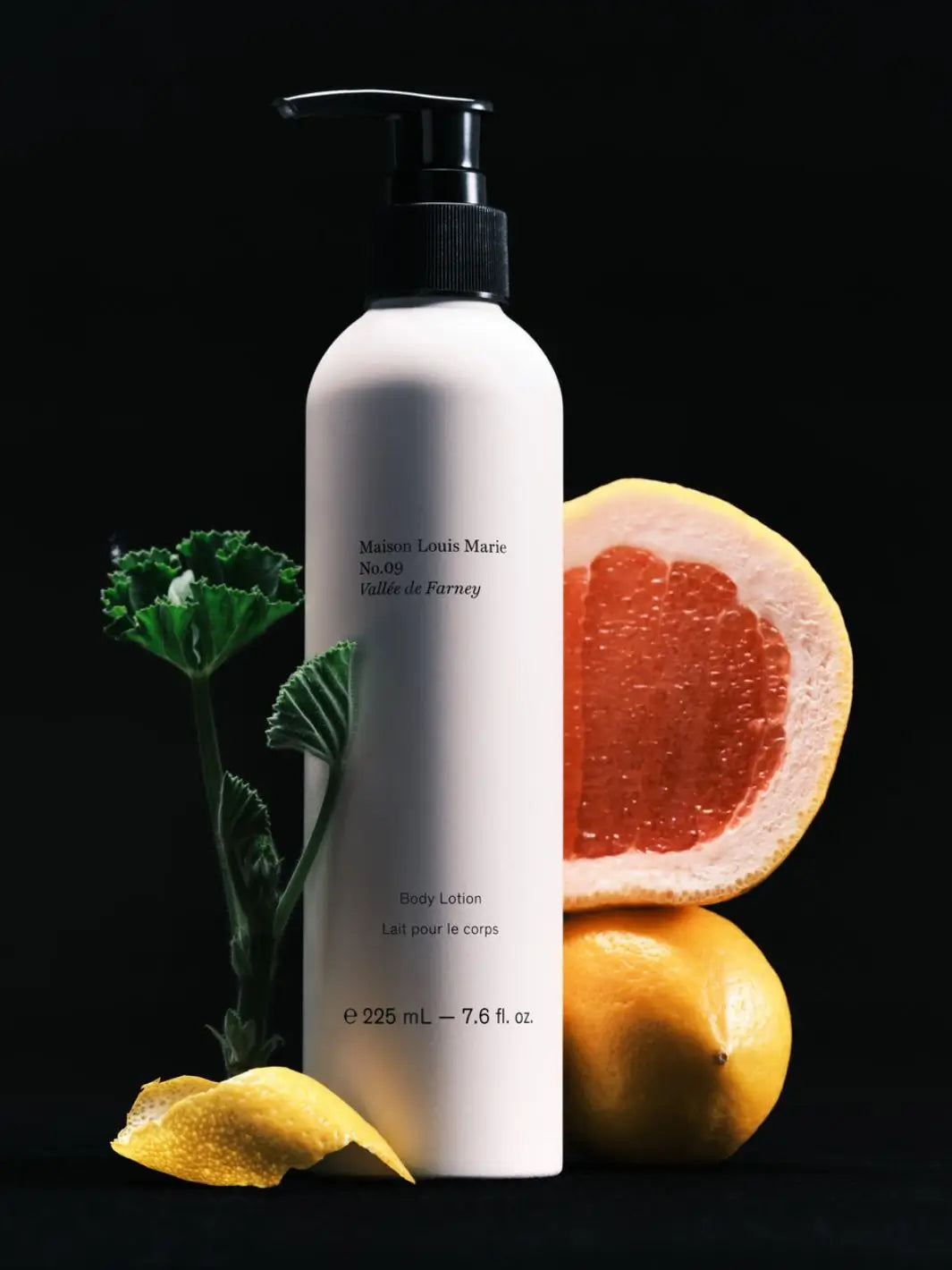 A bottle of Maison Louis Marie No.09 Vallée de Farney Body Lotion, styled with a grapefruit half, a lemon, and green leaves on a solid black background | Twentyseven