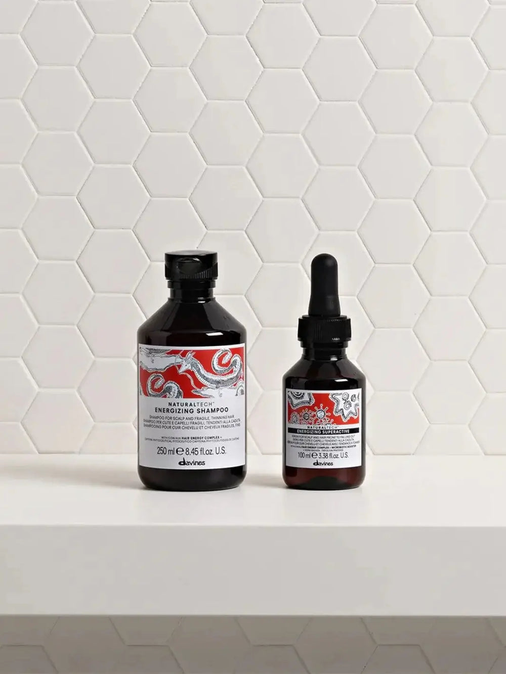 The Davines ENERGIZING Duo, featuring a 250 ml shampoo bottle and a 100 ml dropper serum bottle, stands side-by-side on a white shelf against a white hexagonal tile background | Twentyseven Toronto