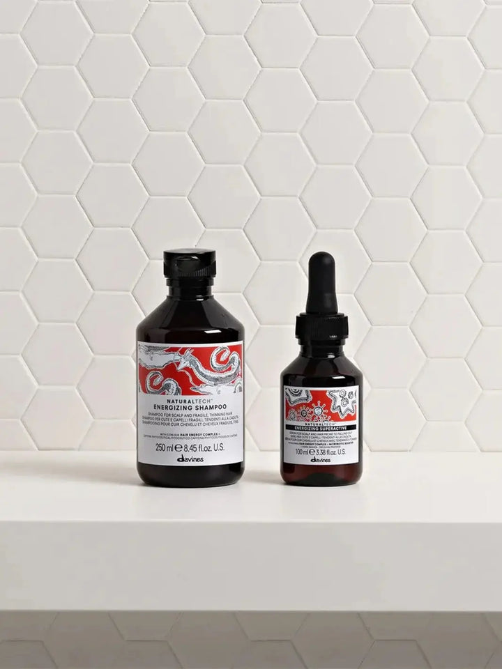 The Davines ENERGIZING Duo, featuring a 250 ml shampoo bottle and a 100 ml dropper serum bottle, stands side-by-side on a white shelf against a white hexagonal tile background | Twentyseven Toronto