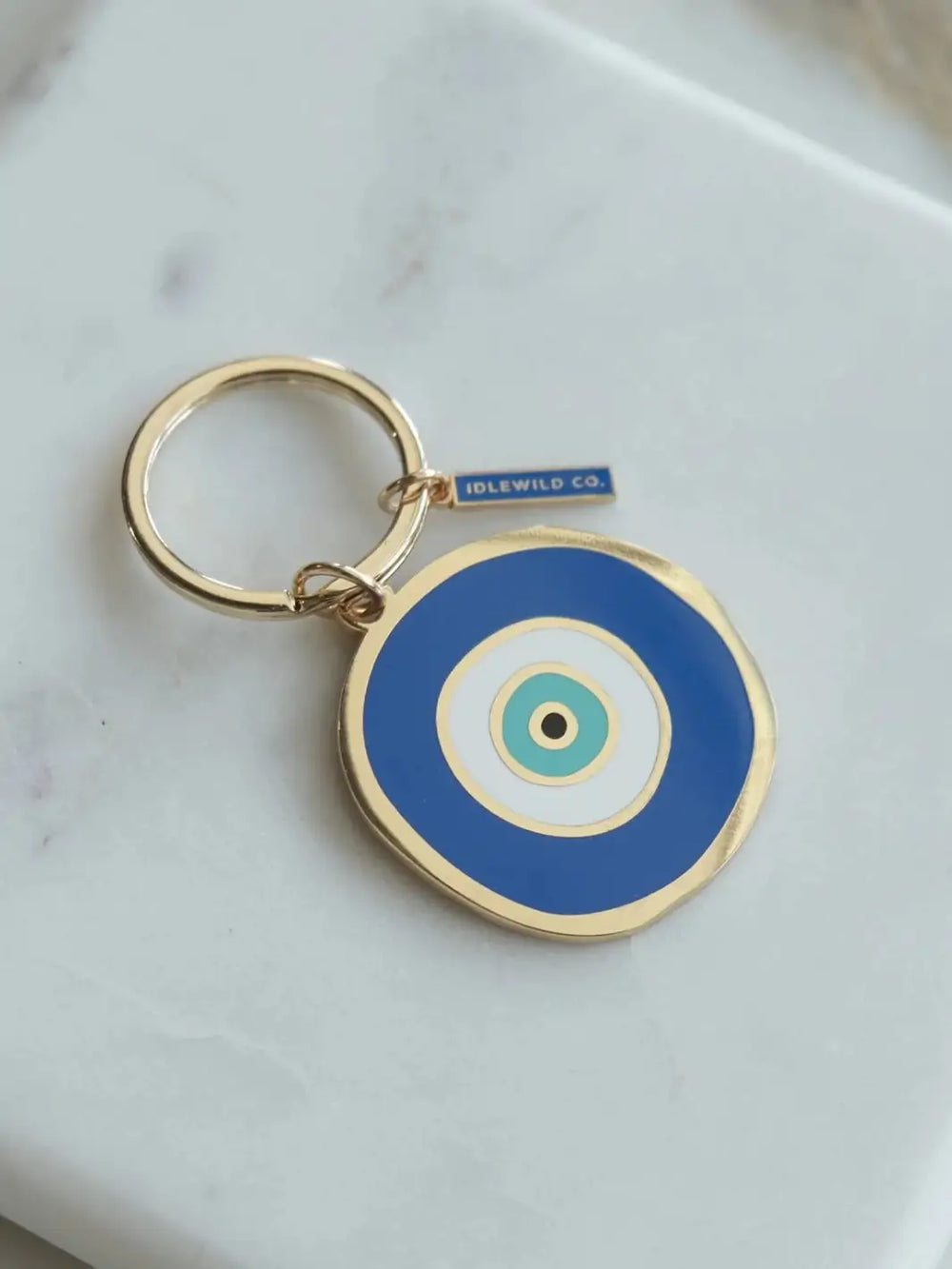 A close-up shot of the blue and gold Idlewild Co. Evil Eye Enamel Keychain lying flat on a white marble surface | Twentyseven