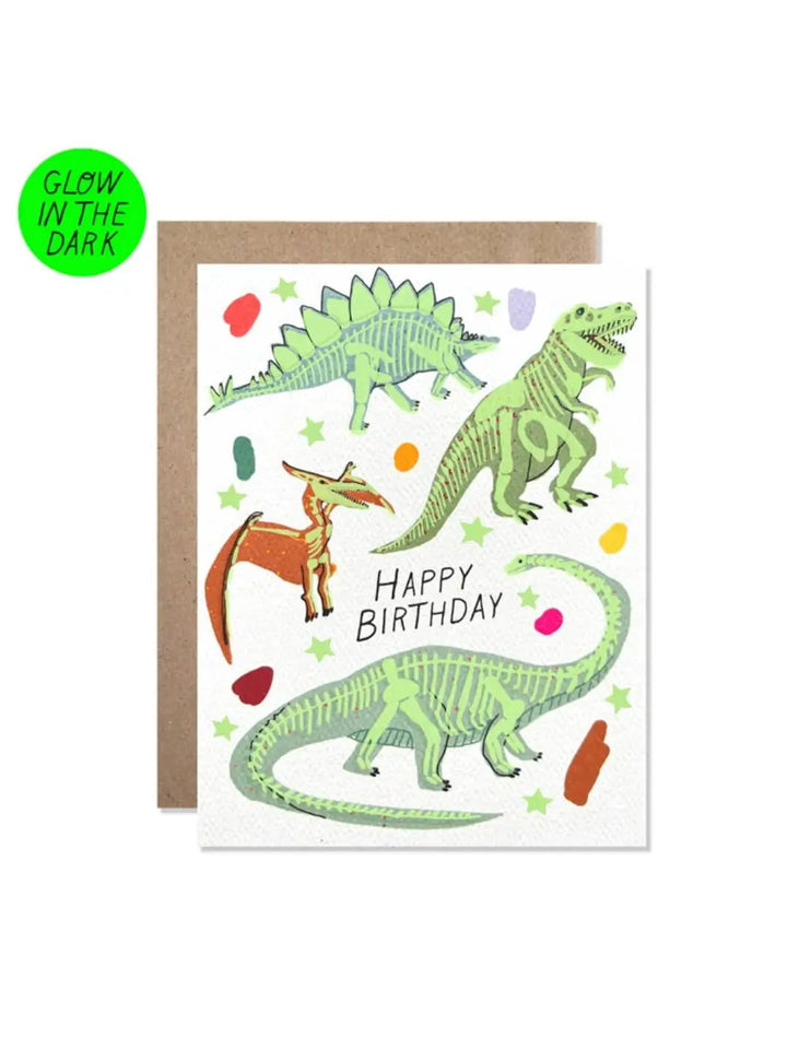 A front view of the Hartland Cards Happy Birthday Glow In The Dark Dinosaurs Card showing the dinosaur illustrations and light-up skeleton details alongside a brown kraft envelope | Twentyseven
