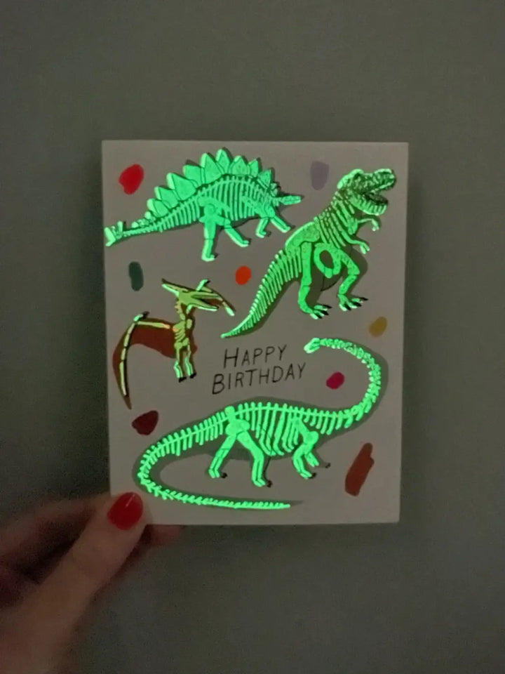 A close-up shot of a hand holding the Hartland Cards Happy Birthday Glow In The Dark Dinosaurs Card in low lighting to demonstrate the glowing skeleton feature | Twentyseven