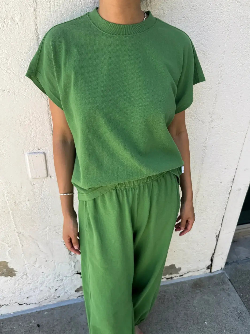 A full-body shot of a model wearing the Le Bon Shoppe Jeanne Tee in Asparagus styled as a monochrome set with matching green pants against a white wall | Twentyseven