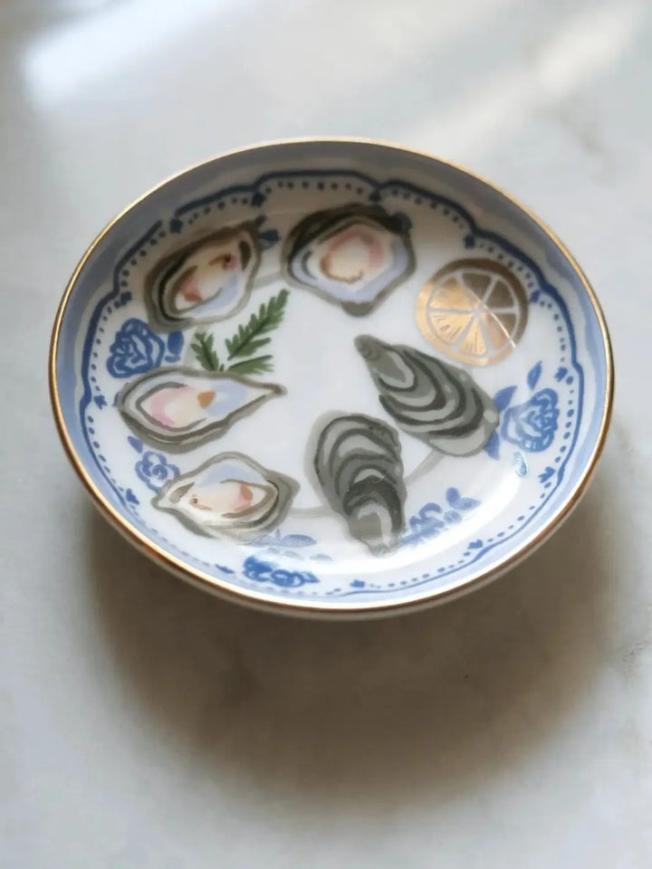 A close-up lifestyle shot resting on a marble surface, featuring the round Idlewild Co. Oysters Porcelain Trinket Dish decorated with an oyster shell platter and gold foil lemon slice illustration | Twentyseven