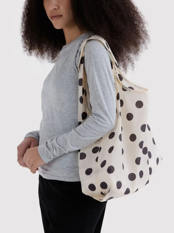 A model with dark curly hair wearing a gray long sleeve and black pants carrying the BAGGU Standard Baggu in Cream and Black Polka Dot over her shoulder | Twentyseven