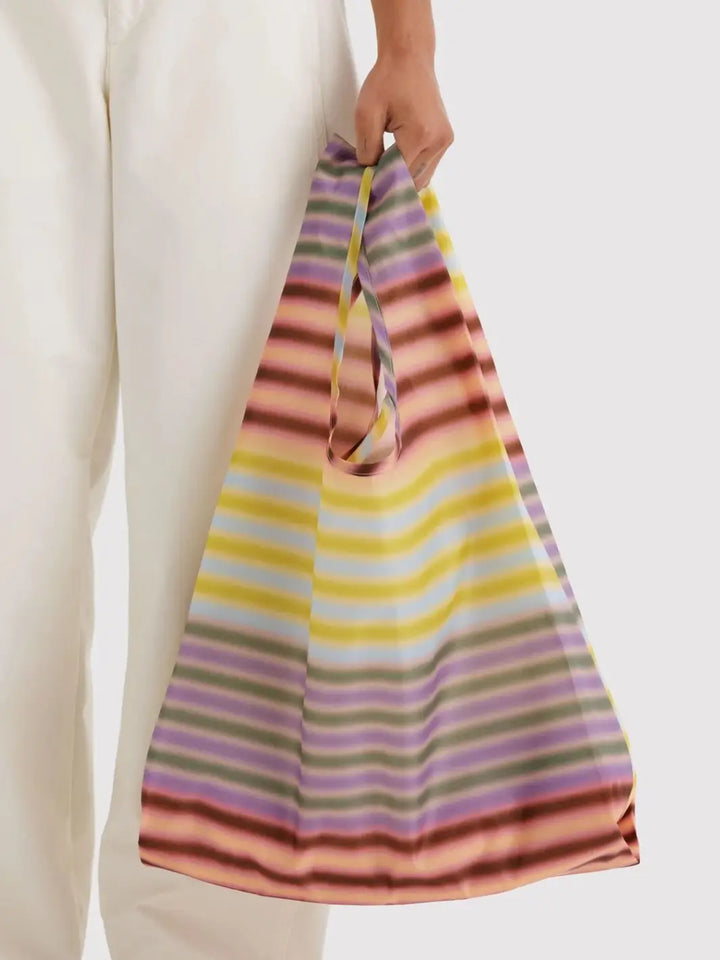 A model wearing white pants holding the BAGGU Standard Baggu in Gradient Stripe Multi by its handles | Twentyseven