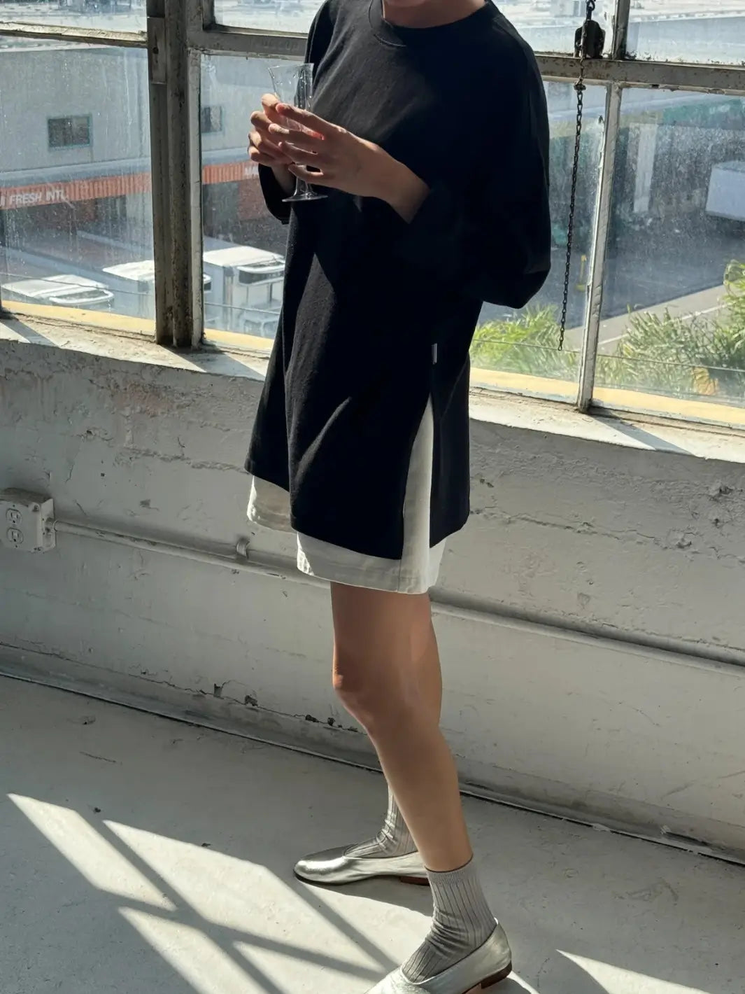 A model standing indoors holding a glass while wearing the long-sleeve Le Bon Shoppe Sunday Tee in Black with side slit detailing | Twentyseven