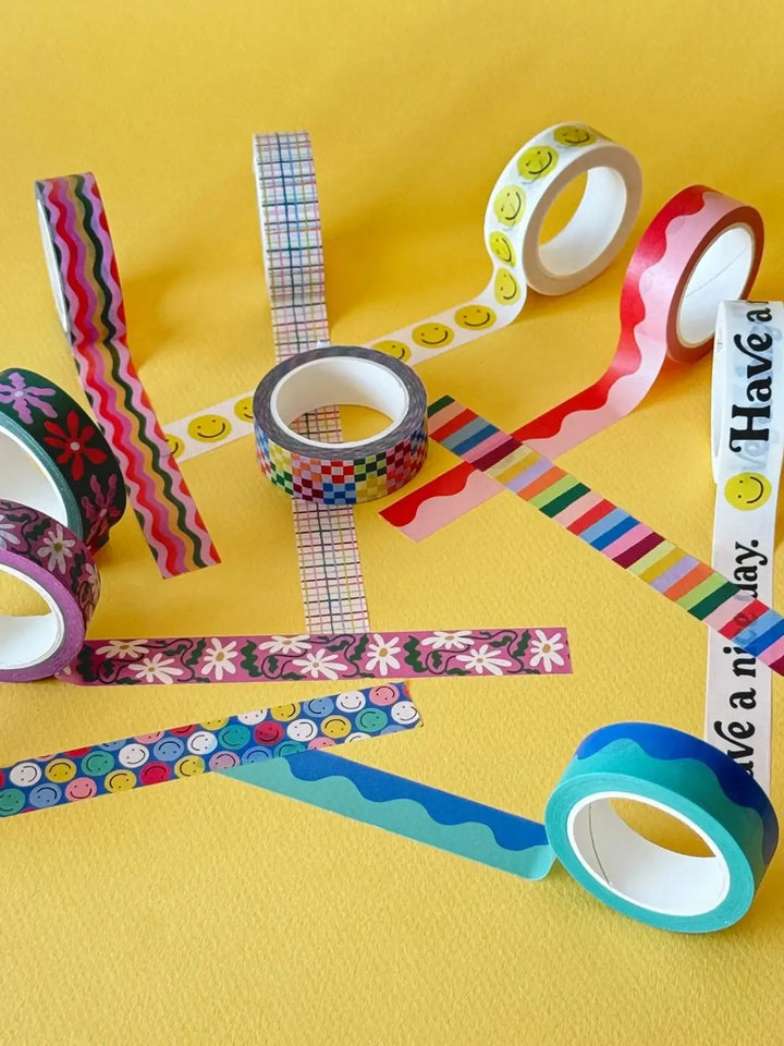 A playful flat lay arrangement featuring strips of the Idlewild Co. Washi Tape, Set of 3 in Rainbow crisscrossing a bright yellow background among other colorful, decorative tape rolls | Twentyseven