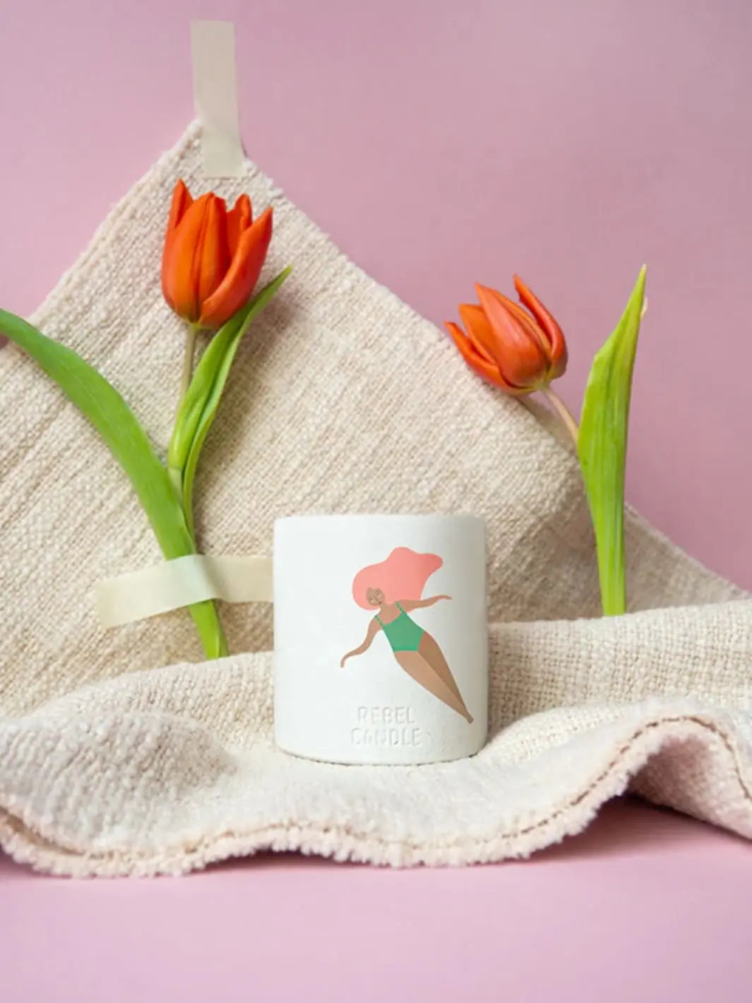 Maison Matine A Contre Courant Scented Candle with a design of a woman in a green swimsuit and red shoes, placed on a textured towel with orange tulips 160g | Twentyseven Toronto