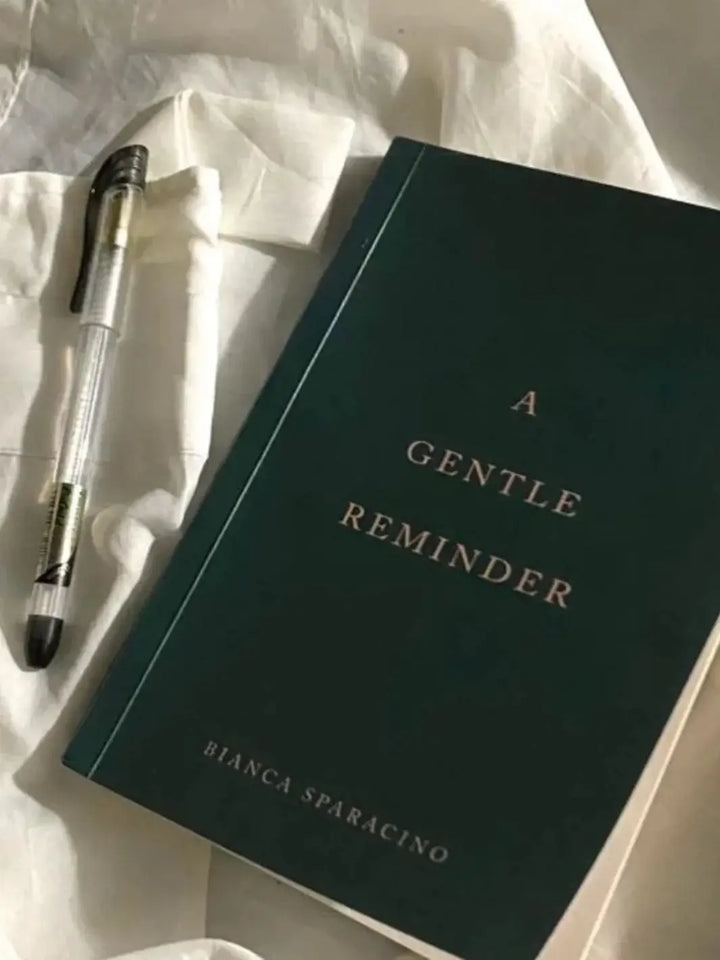 The book titled 'A Gentle Reminder' by Bianca Sparacino rests on rumpled white fabric next to a clear plastic pen | Twentyseven Toronto