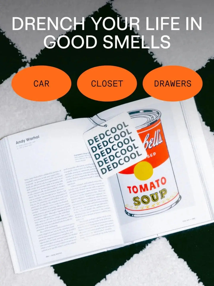 A square Dedcool Air Freshener 01 "Taunt" tag rests on an open Andy Warhol art book against a black and white checkered background with orange promotional text bubbles | Twentyseven Toronto