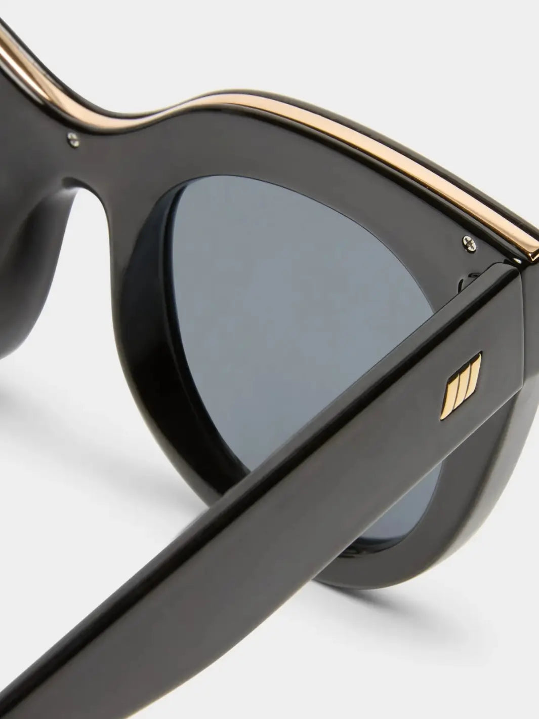A macro close-up highlighting the metallic gold strip along the top frame and the small gold emblem on the temple of the Le Specs Air Heart - Black sunglasses | Twentyseven Toronto