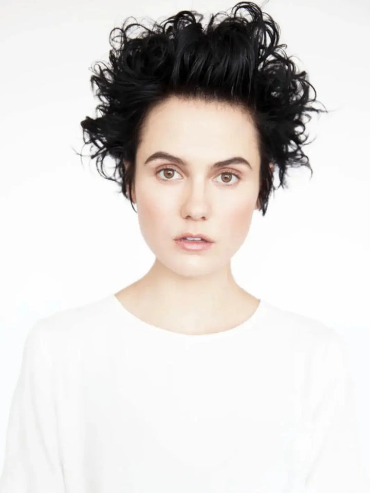 A front-facing headshot of a model showcasing short, textured, and voluminous dark curls styled with R+Co AIRCRAFT Pomade Mousse | Twentyseven Toronto