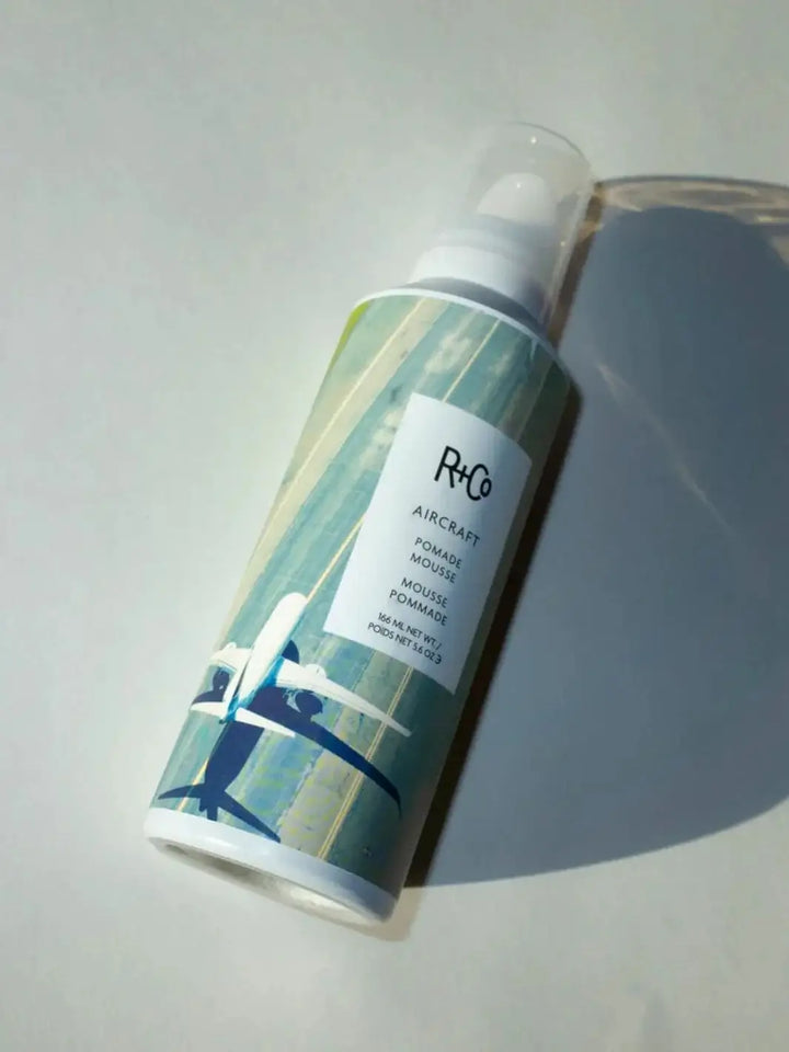 An aerosol bottle of R+Co AIRCRAFT Pomade Mousse lies diagonally on a white surface, casting a distinct shadow | Twentyseven Toronto