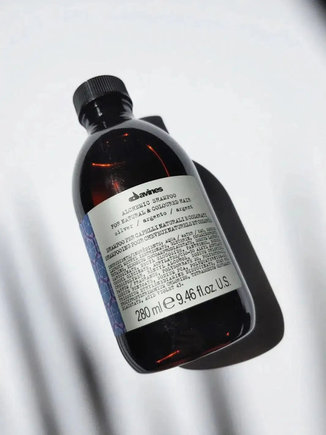 An amber plastic bottle of Davines ALCHEMIC Silver Shampoo rests at an angle on a clean white surface with shadows | Twentyseven Toronto