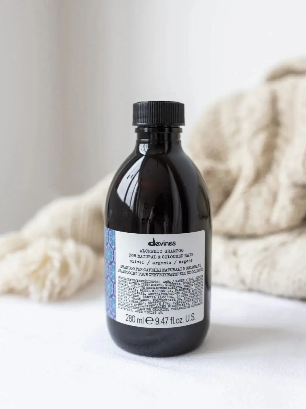 A bottle of Davines ALCHEMIC Silver Shampoo stands on a white surface with a cream-colored knit blanket blurred in the background | Twentyseven Toronto