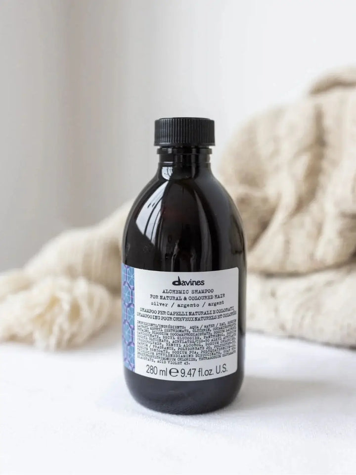 A bottle of Davines ALCHEMIC Silver Shampoo stands on a white surface with a cream-colored knit blanket blurred in the background | Twentyseven Toronto