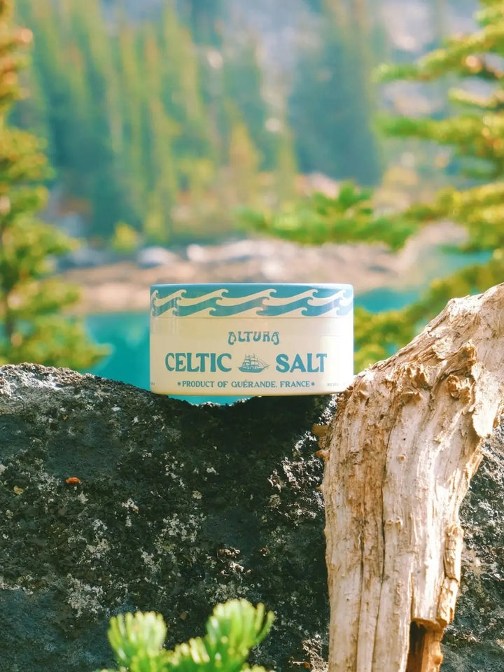 A lifestyle shot shows a container of Altura Celtic Sea Salt resting on a dark rock with a scenic, out-of-focus background of a turquoise mountain lake and evergreen trees | Twentyseven Toronto