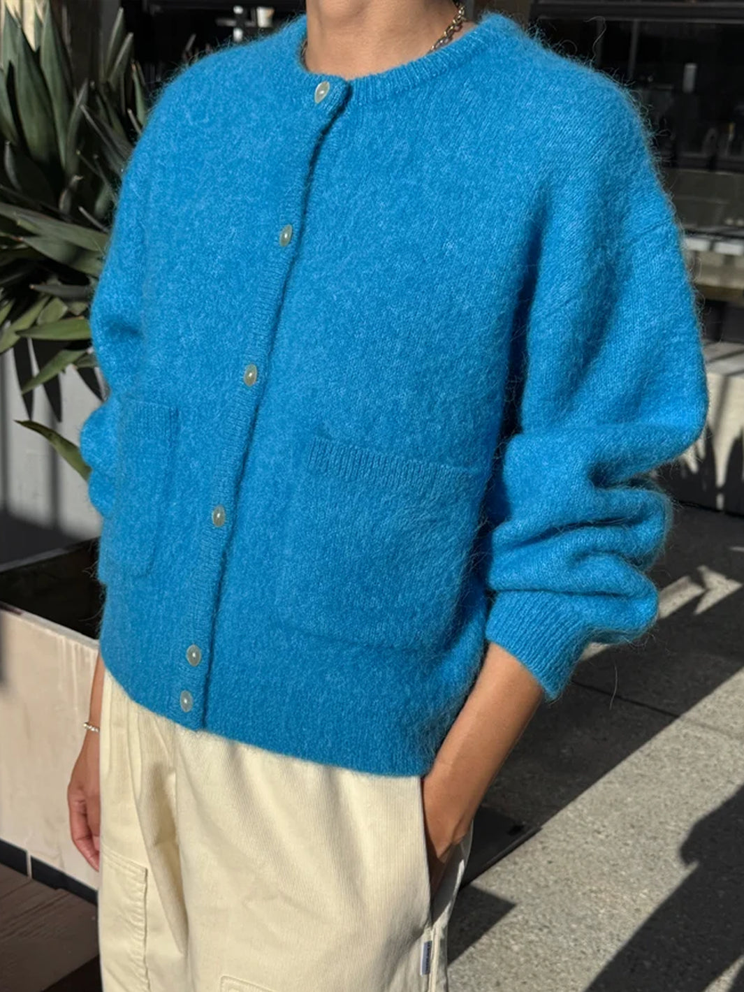 Angled view of the Le Bon Shoppe Amelie Cardigan in turquoise blue, styled with cream trousers, highlighting its relaxed silhouette, ribbed cuffs and button-front design | Twentyseven Toronto