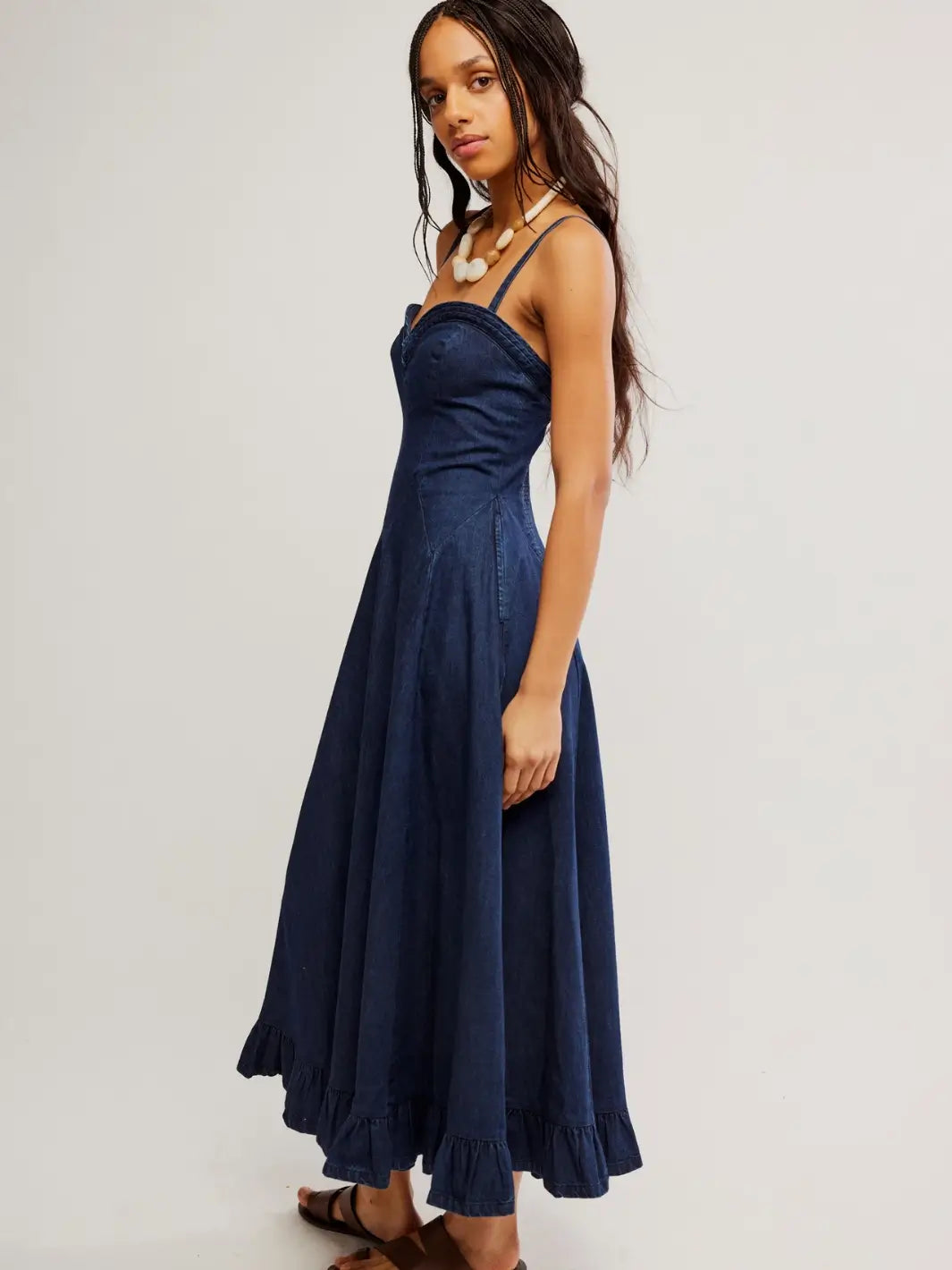 A side three-quarter view of a model wearing the Free People Another Season Denim Midi Dress in Rinse, showcasing its ruffled hemline and dark wash denim fabric | Twentyseven Toronto