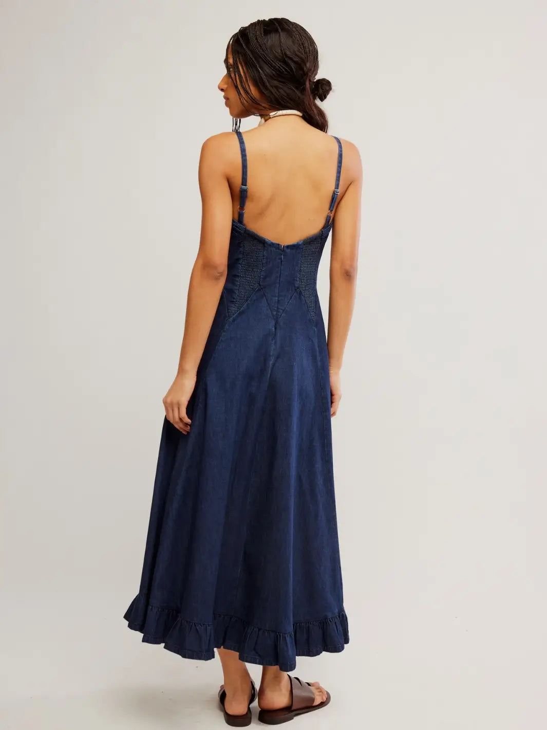A rear view of a model highlighting the low-cut back design, adjustable shoulder straps, and smocked panels of the Free People Another Season Denim Midi Dress in Rinse | Twentyseven Toronto