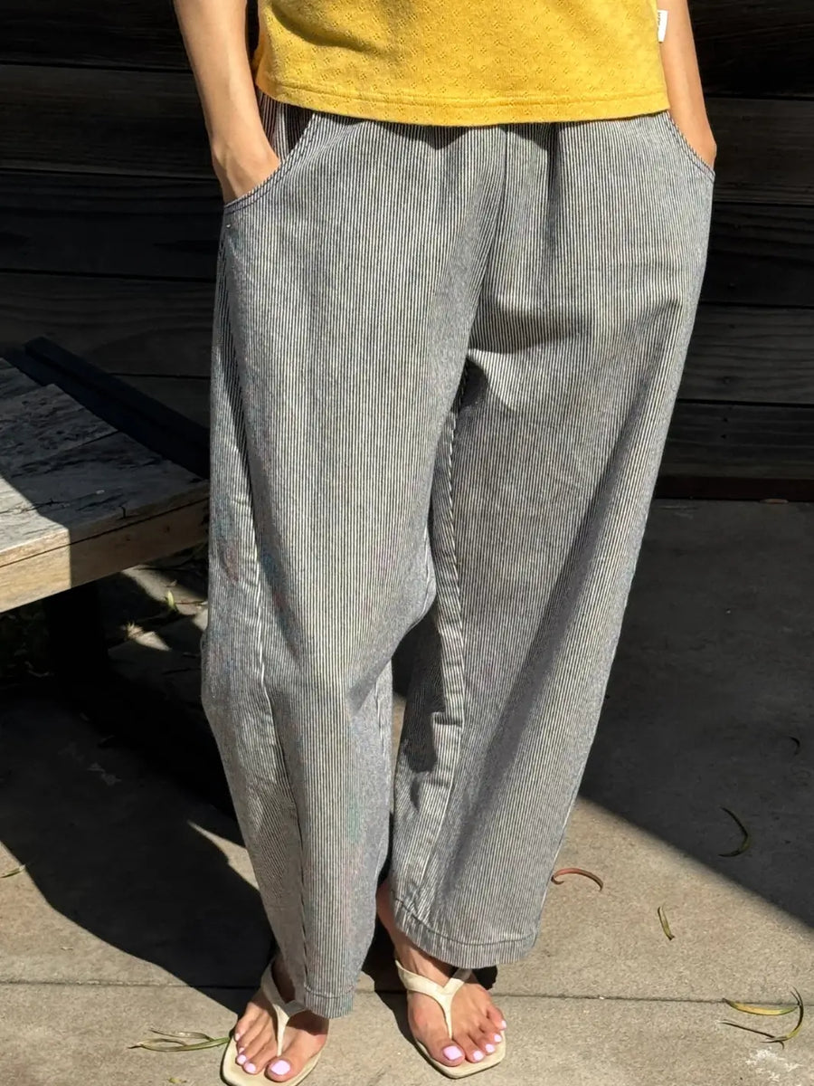 An outdoor shot shows a person standing with their hands in their pockets, wearing the slouchy, vertically striped Le Bon Shoppe Arc Pants in Blue Stripe paired with a mustard yellow top and white sandals | Twentyseven Toronto