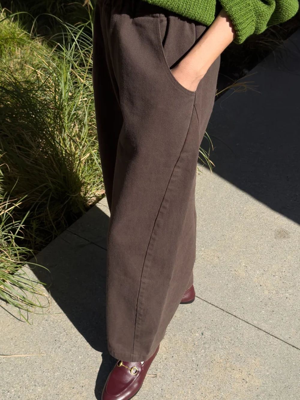Side view of umber brown Arc Pants featuring a relaxed wide-leg silhouette and side pockets, styled with a green sweater and burgundy loafers | Twentyseven Toronto