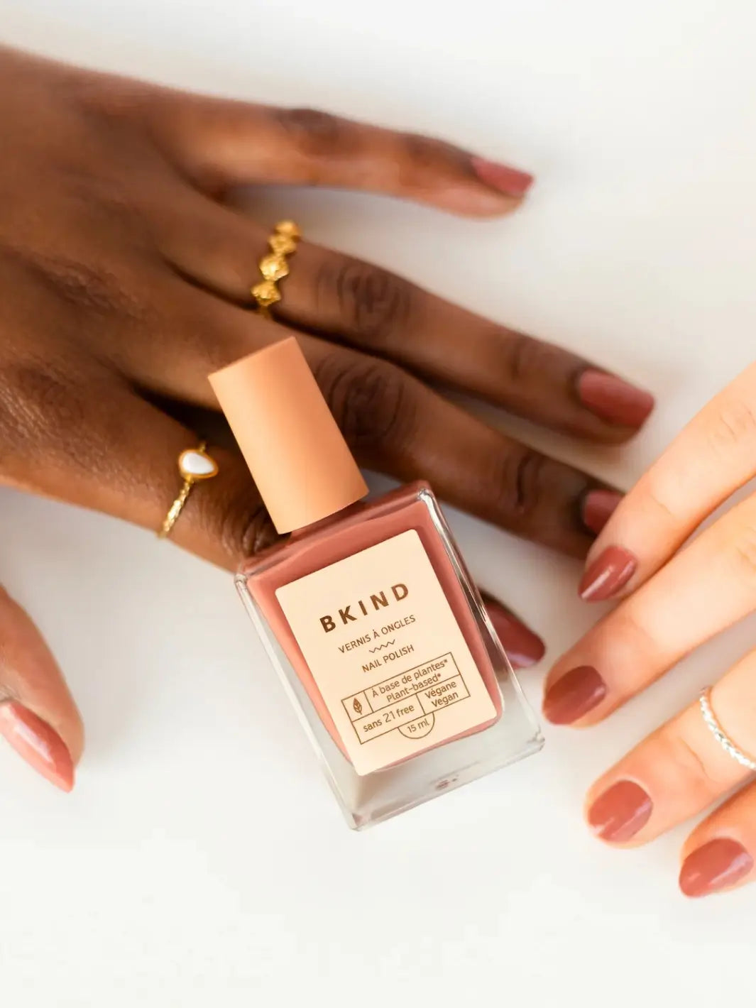 A bottle of BKIND Nail Polish in Arizona sits between two hands with different skin tones, both showing manicured nails painted in the same dusty rose shade | Twentyseven Toronto