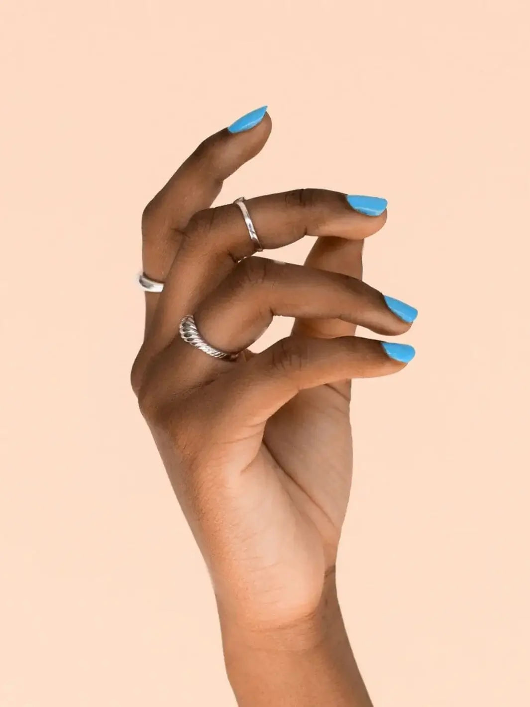 A dark-skinned hand with silver rings showcases a neat, glossy manicure using BKIND Nail Polish in Blue Lagoon against a peach background | Twentyseven Toronto