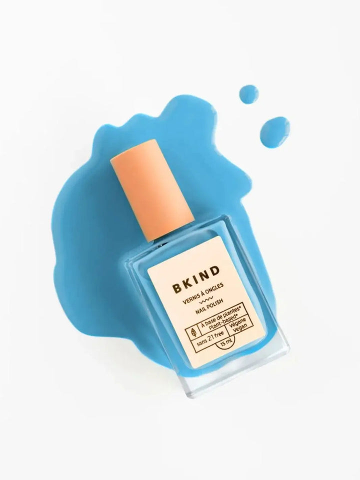 A square bottle of BKIND Nail Polish in Blue Lagoon sits atop a vibrant splash and droplets of the matching blue polish liquid on a white background | Twentyseven Toronto