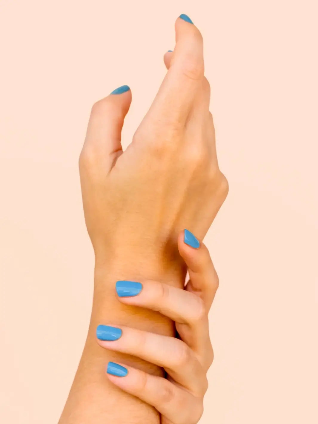 A close-up shot of a hand gently clasping the opposite wrist, showcasing a glossy sky-blue manicure painted with BKIND Nail Polish in Blue Lagoon against a peach background | Twentyseven Toronto