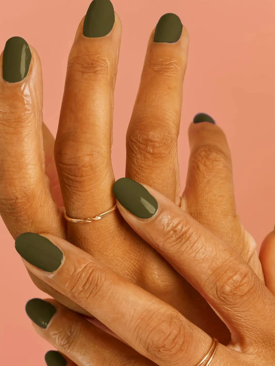 A close-up of a medium-skinned hand wearing thin gold rings and showcasing an olive green manicure painted with BKIND Nail Polish in La Route Verte against a pink background | Twentyseven Toronto
