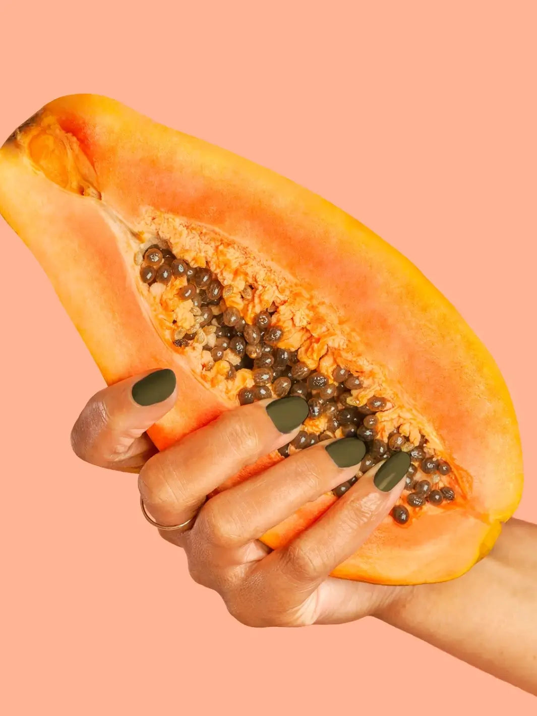 A hand with a glossy olive green manicure holds a sliced open, seed-filled papaya, showcasing the shade of BKIND Nail Polish in La Route Verte against a coral background | Twentyseven Toronto