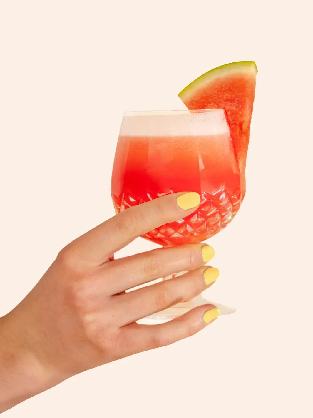 A hand with a bright yellow manicure holds a textured glass filled with a pink cocktail and garnished with a watermelon slice, showcasing the BKIND Nail Polish in Tulum | Twentyseven Toronto