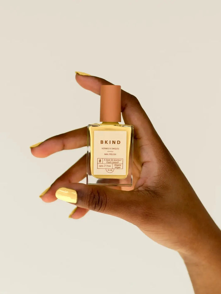 A close-up of a dark-skinned hand holding a square bottle of BKIND Nail Polish in Tulum, displaying fingernails painted in the same vibrant, pastel yellow shade | Twentyseven Toronto
