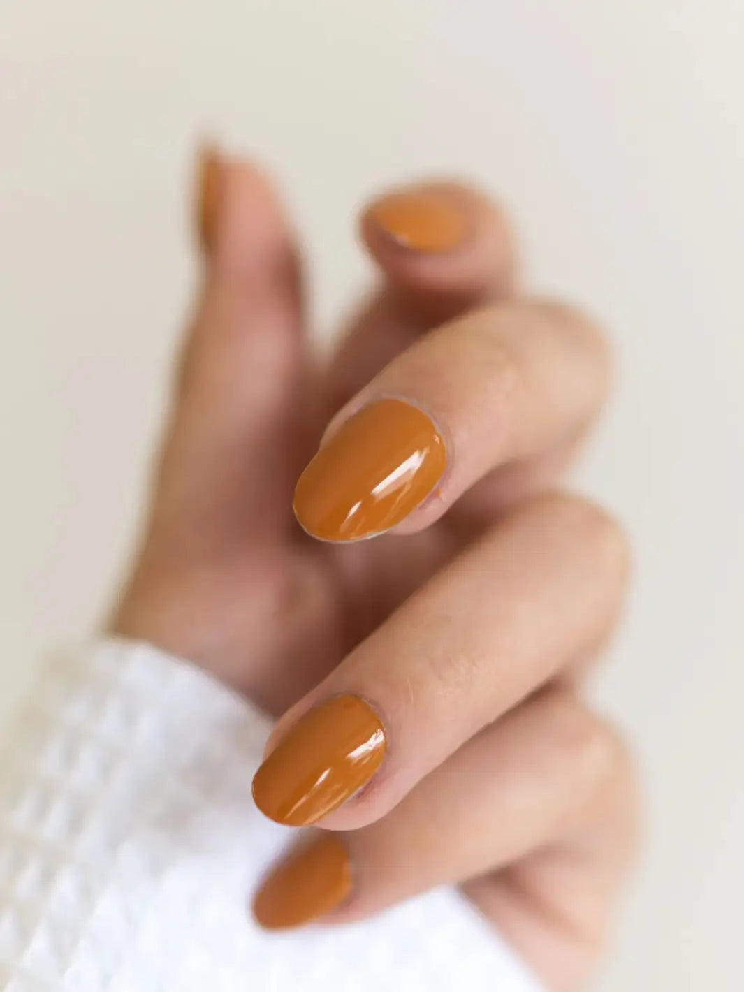 A close-up shot of a hand wearing a textured white sleeve, showcasing a glossy, warm mustard-brown manicure with BKIND Nail Polish in Val-d'Or against a blurred neutral background | Twentyseven Toronto