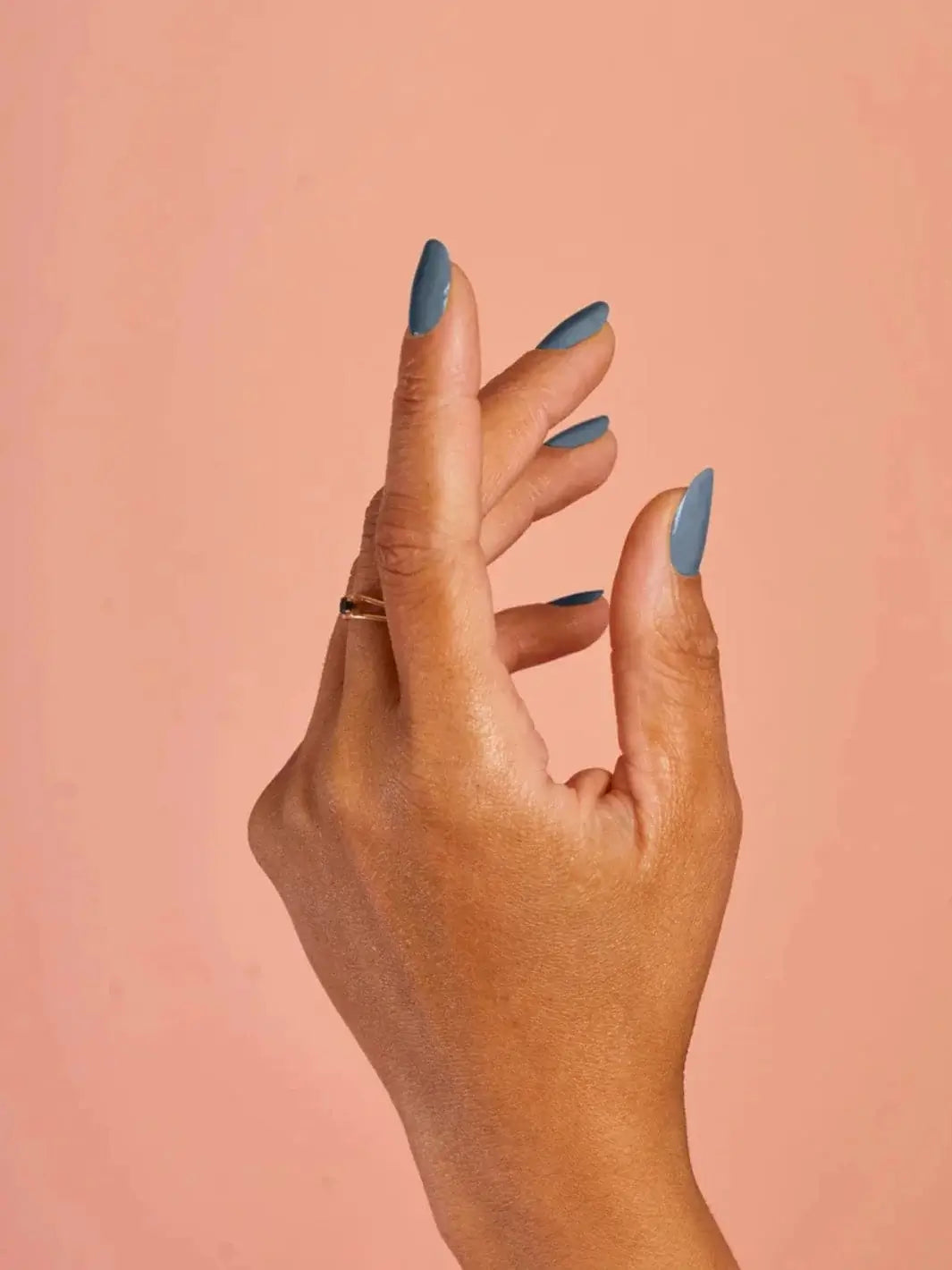 A hand wearing a gold ring showcases a glossy, slate-green manicure with BKIND Nail Polish in Verdun Beach against a solid pink background | Twentyseven Toronto