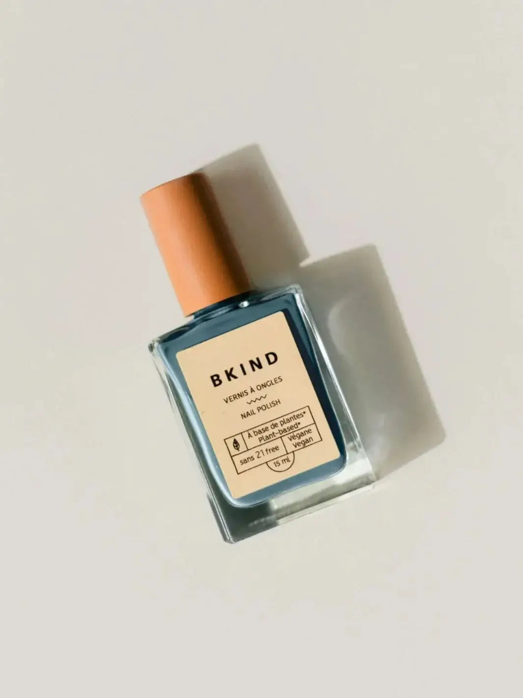 A square glass bottle of BKIND Nail Polish in Verdun Beach with a light beige cap rests diagonally against a clean, off-white background | Twentyseven Toronto