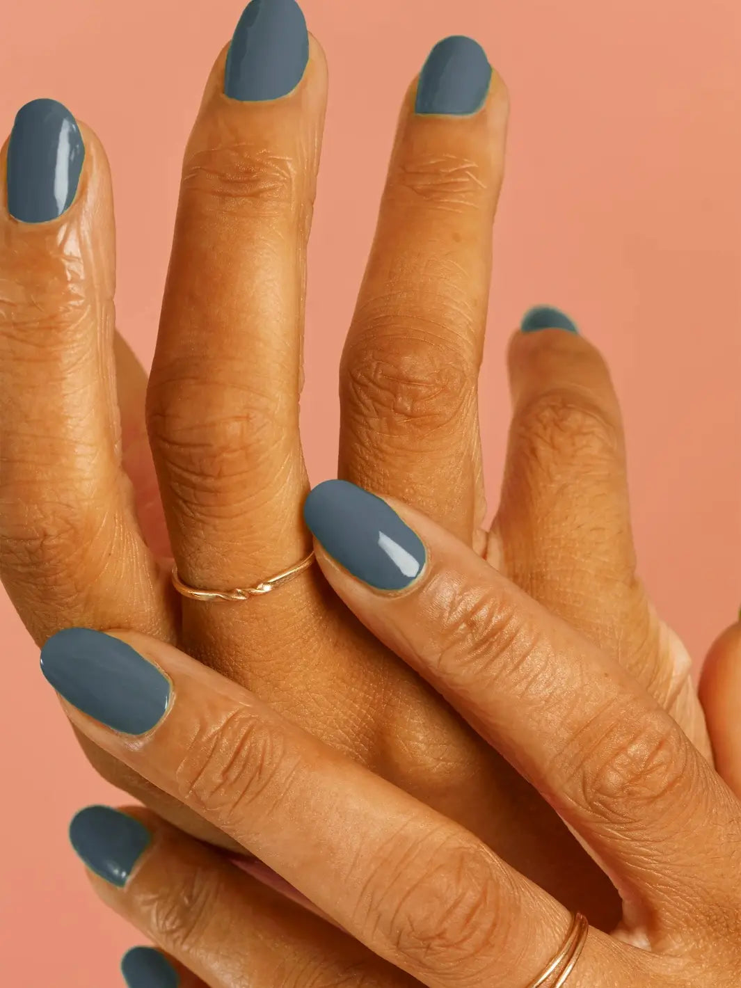 A close-up of a hand wearing thin gold rings and showcasing a glossy, muted green manicure painted with BKIND Nail Polish in Verdun Beach against a pink background | Twentyseven Toronto